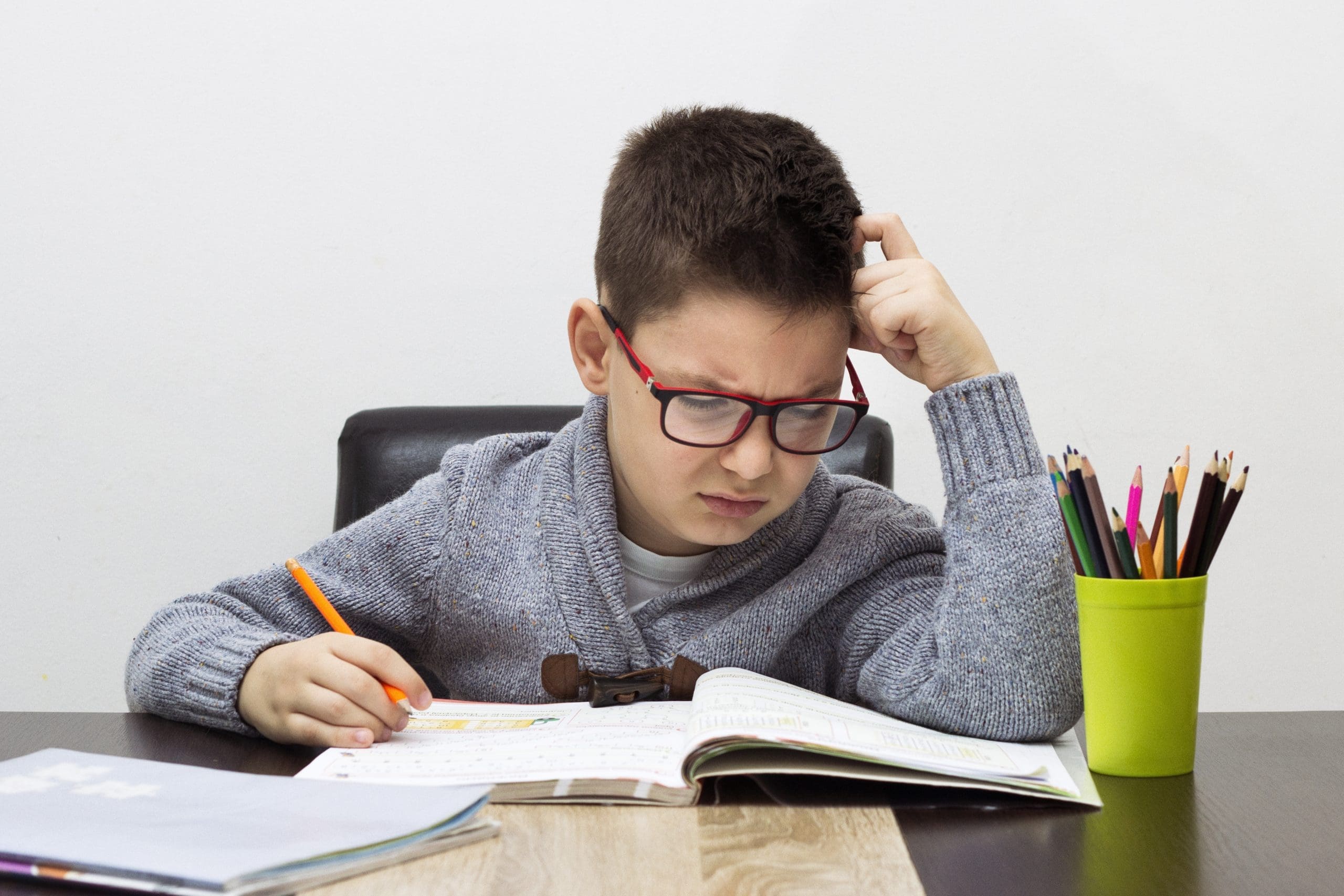 Boy in gray sweater studying, holding pencil, colorful pencils in cup.