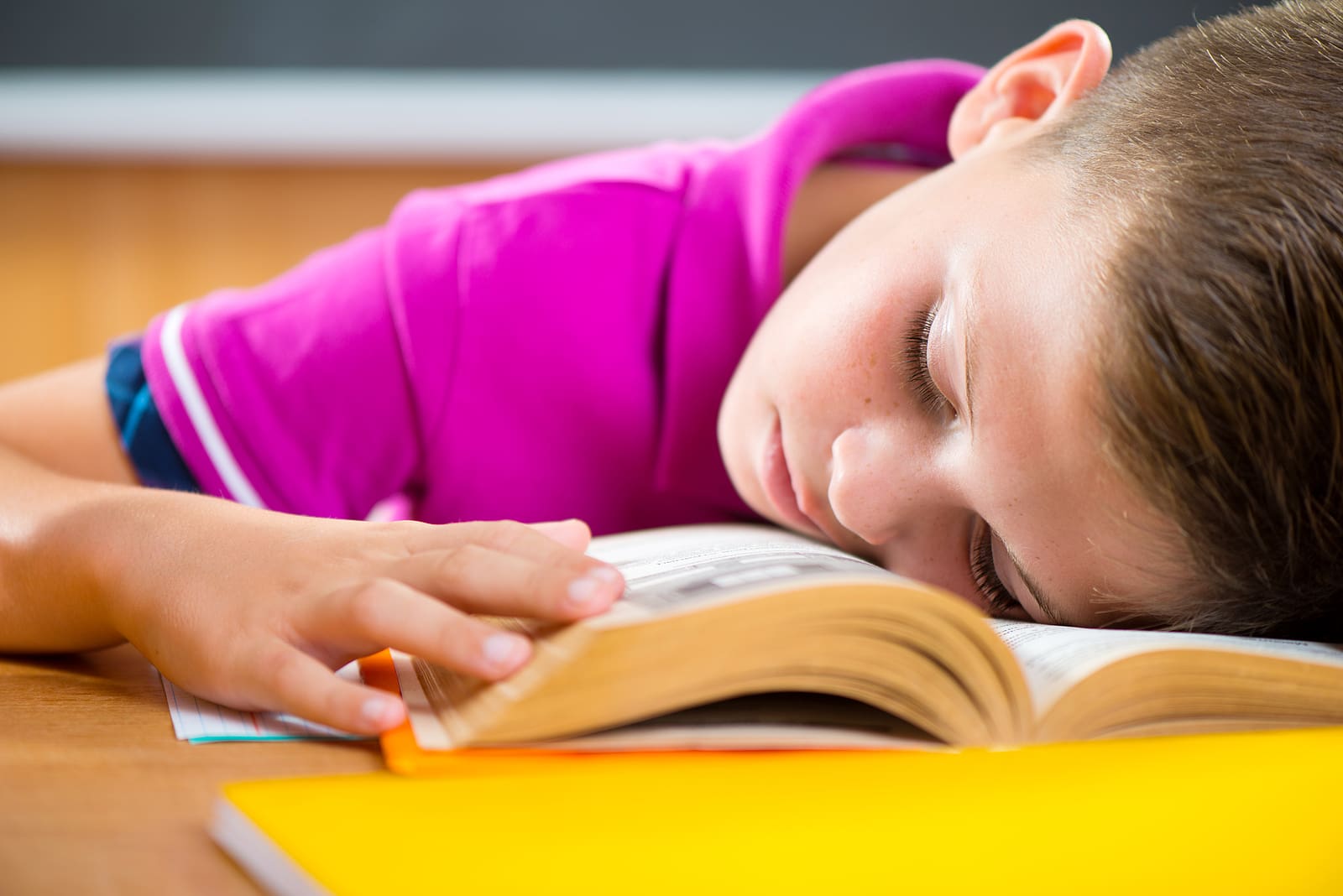 Boy in pink shirt sleeping on open book, yellow notebook visible.