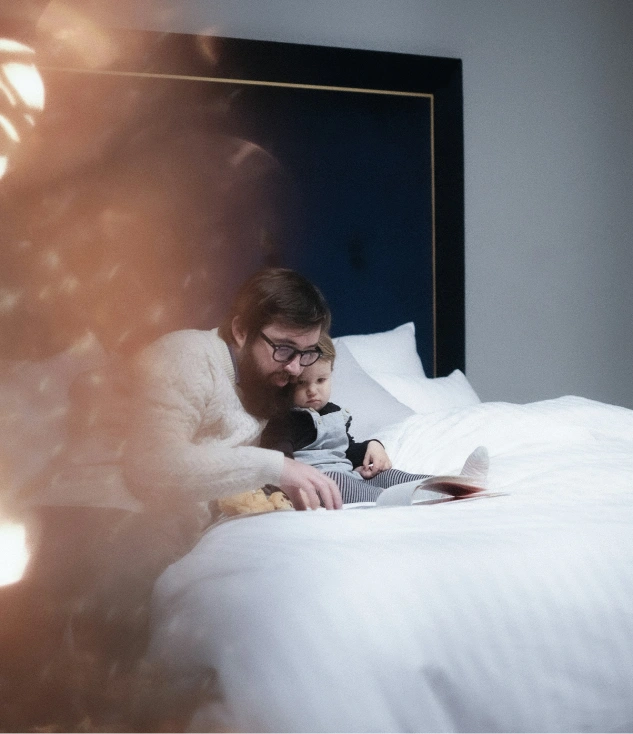 Man and child reading on bed, cozy lighting, photo, no text visible.