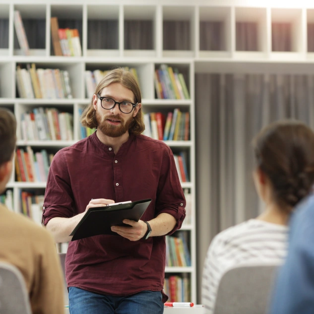 Man in maroon shirt holding clipboard, teaching in a library setting.