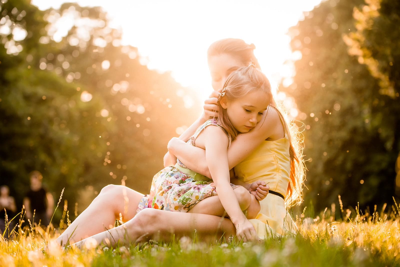Mother and daughter hugging in sunlit park, wearing floral and yellow dresses.