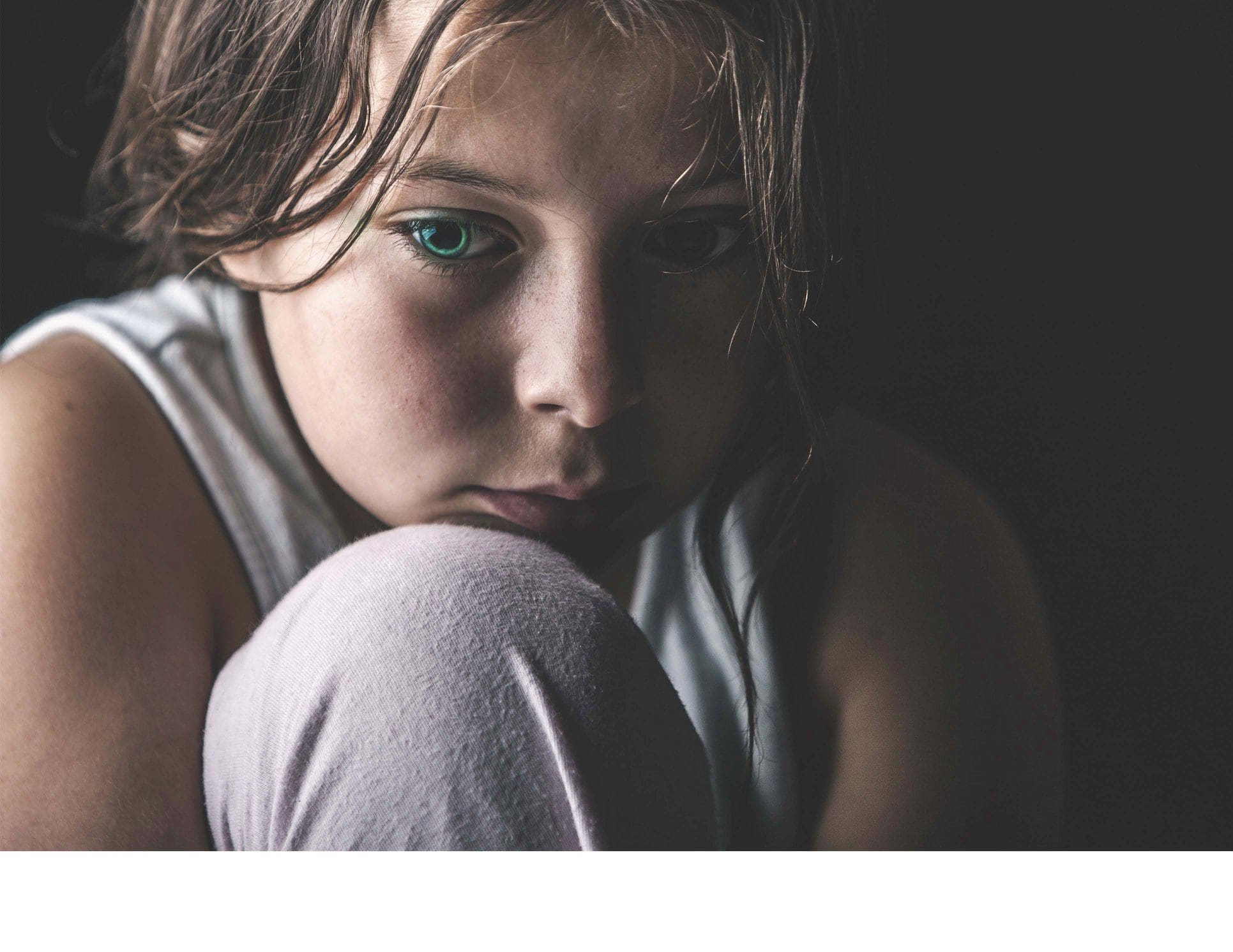 Young girl with green eyes, sitting pensively, in dimly lit photograph.
