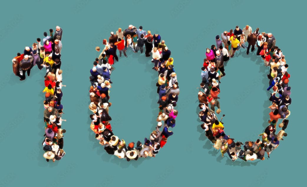 Crowd forming "100" on teal background, colorful clothing, aerial photo.